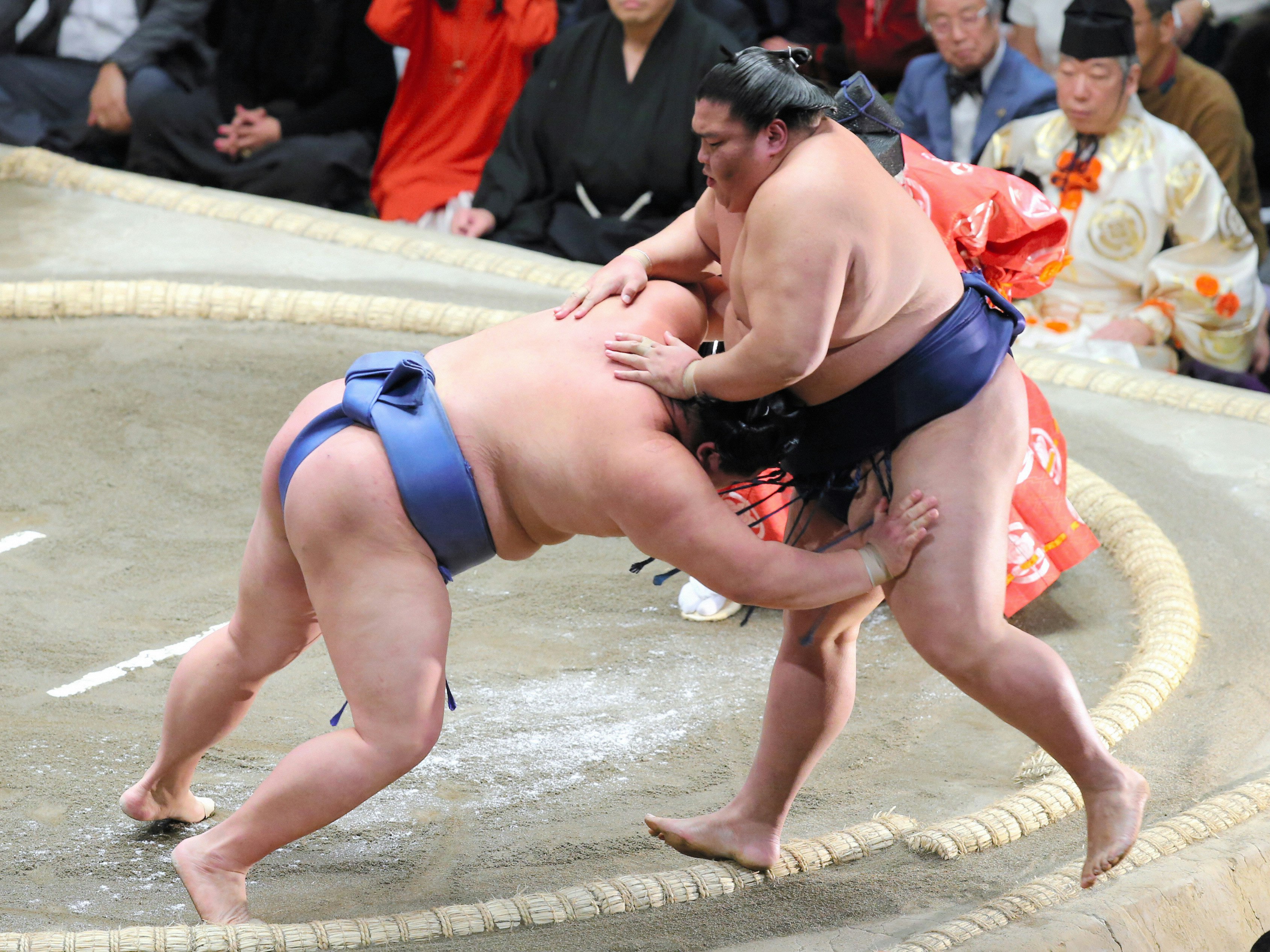 In a dramatic moment in a wrestling ring, circled by salt, one sumo wrestler bends forward to lunge at his opponent, who resists, with a look of concentration